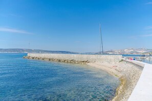 Ein Küstenweg mit einer Steinmauer, klarem, blauem Wasser und sanften Hügeln in der Ferne.