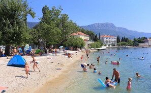 A beach with people swimming and sunbathing, tents, and buildings in the background. - Apartments Castel L (Kastela)