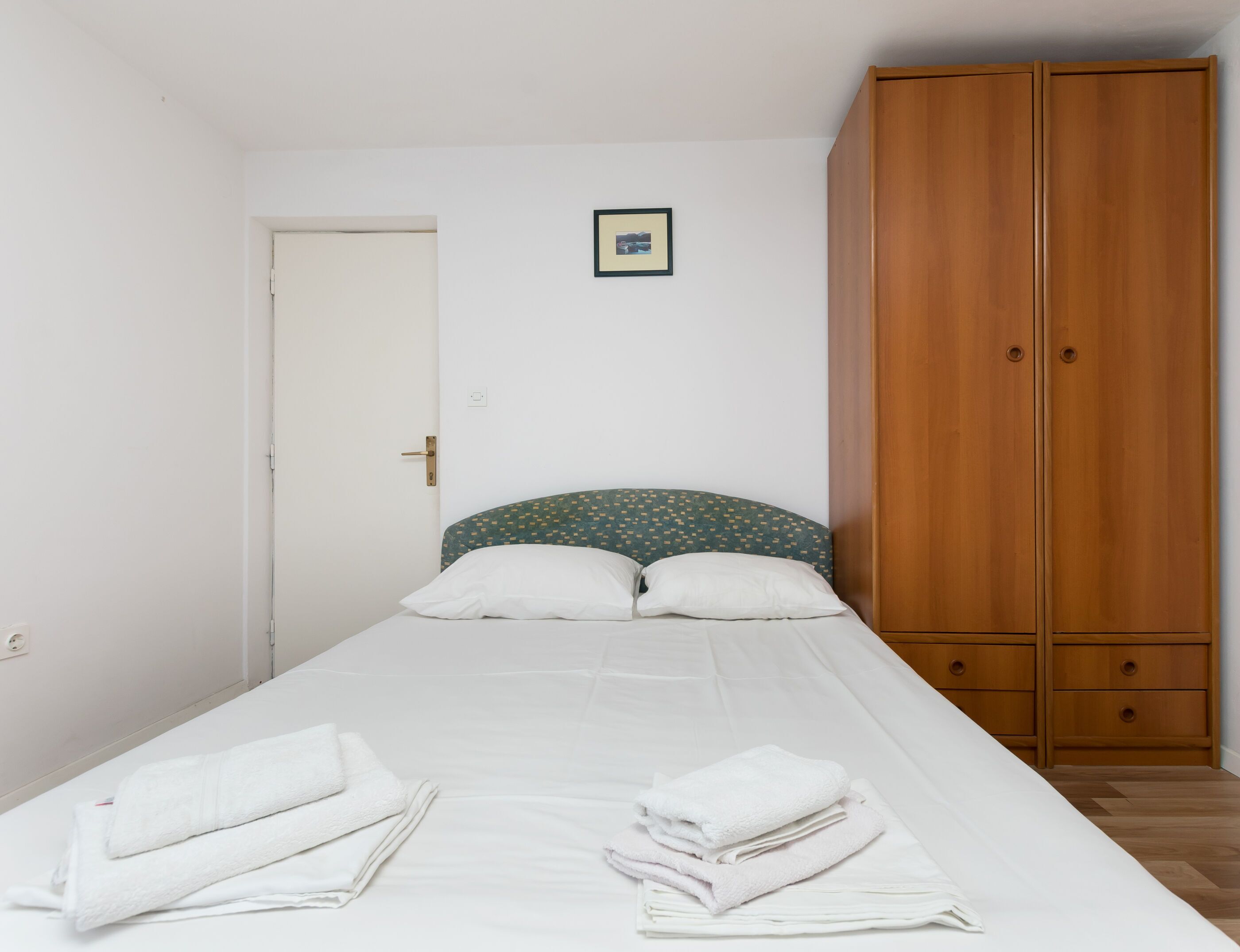 Un lit simple avec du linge de lit blanc et deux oreillers, une armoire en bois et une porte sur laquelle est accrochée une petite photo encadrée.