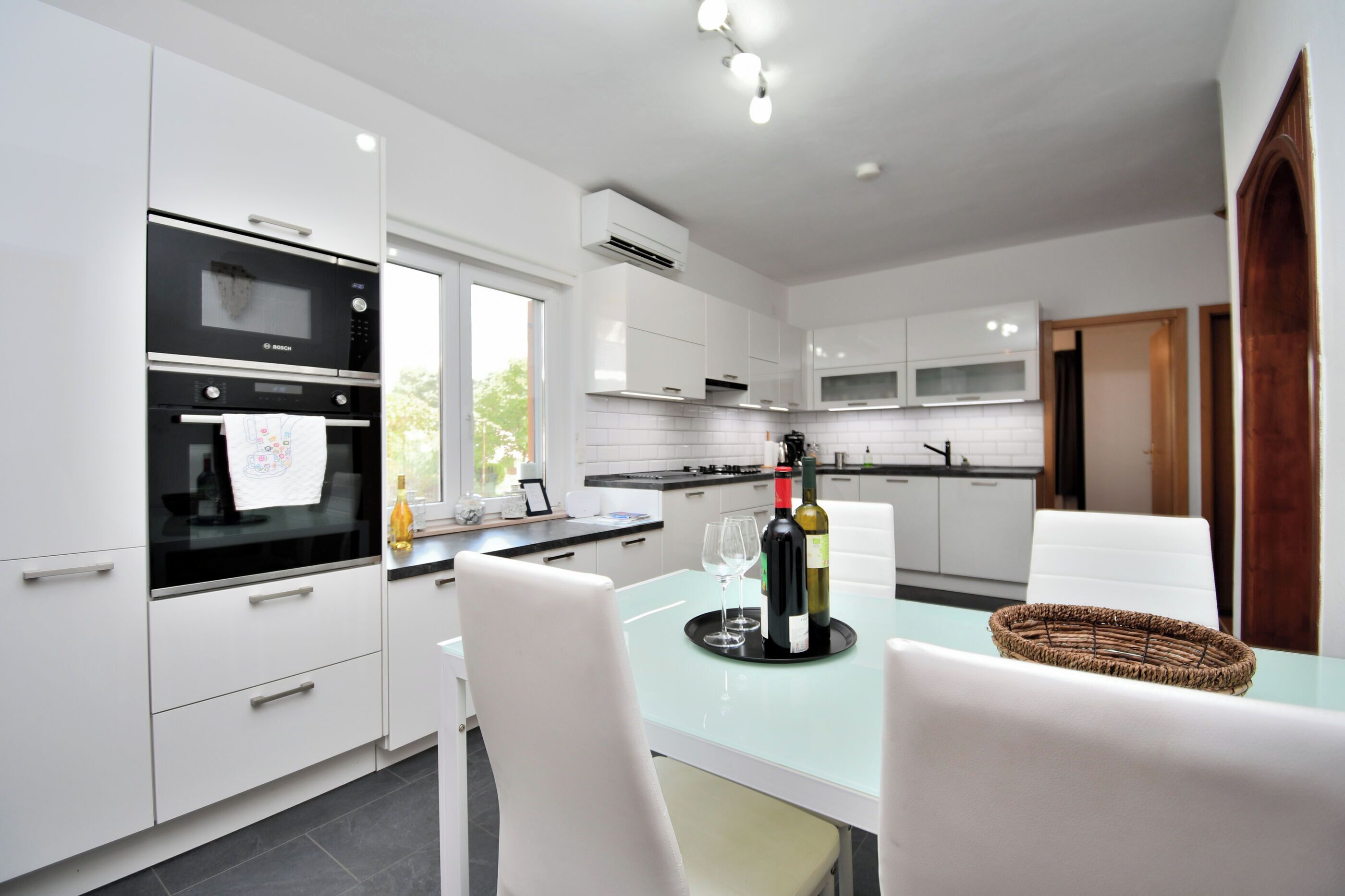 A modern kitchen with white cabinetry, a built-in oven, and a dining area with a glass table.