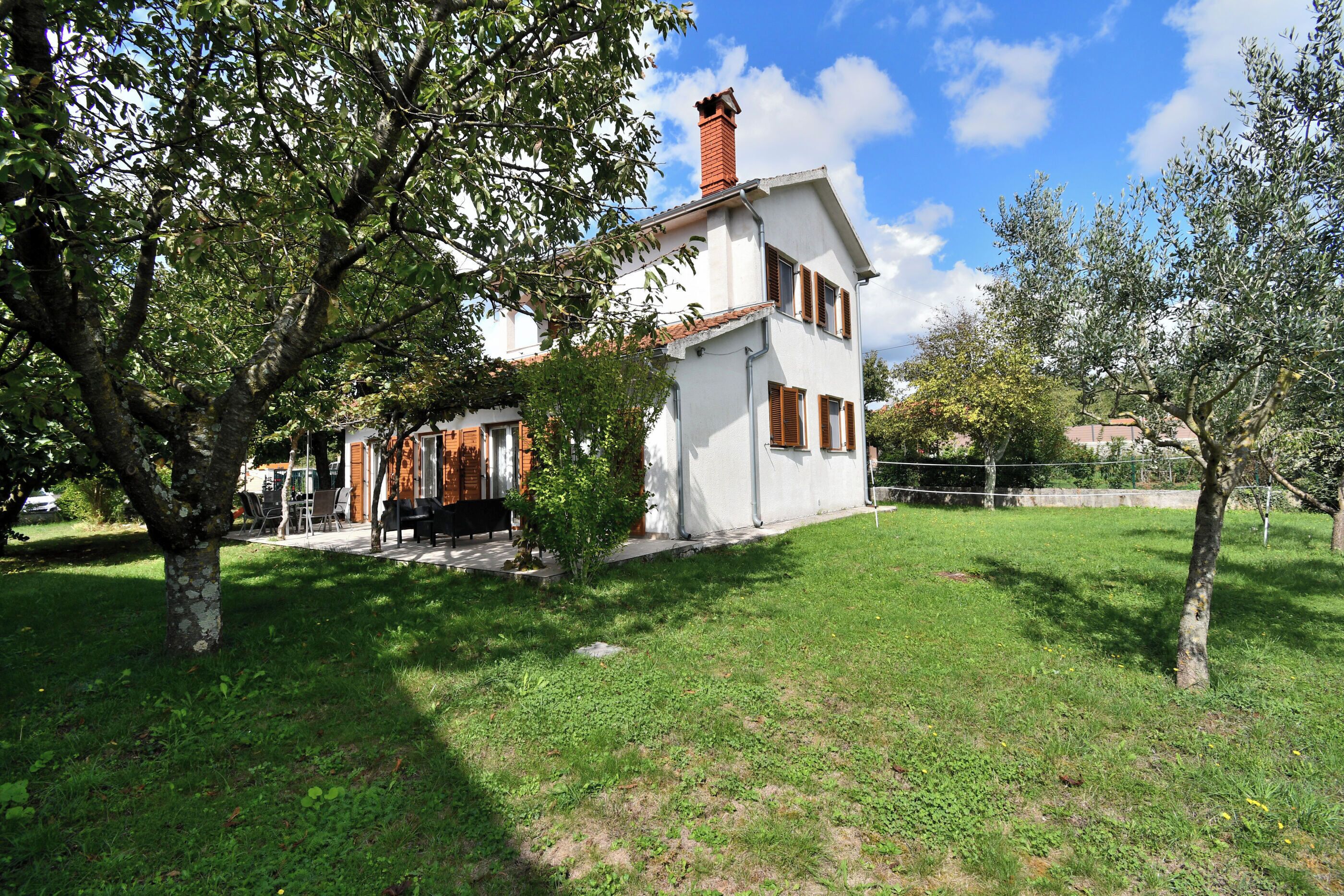 Une maison à deux étages, à l’extérieur blanc et aux volets bruns, entourée d’une pelouse bien entretenue et d’arbres.