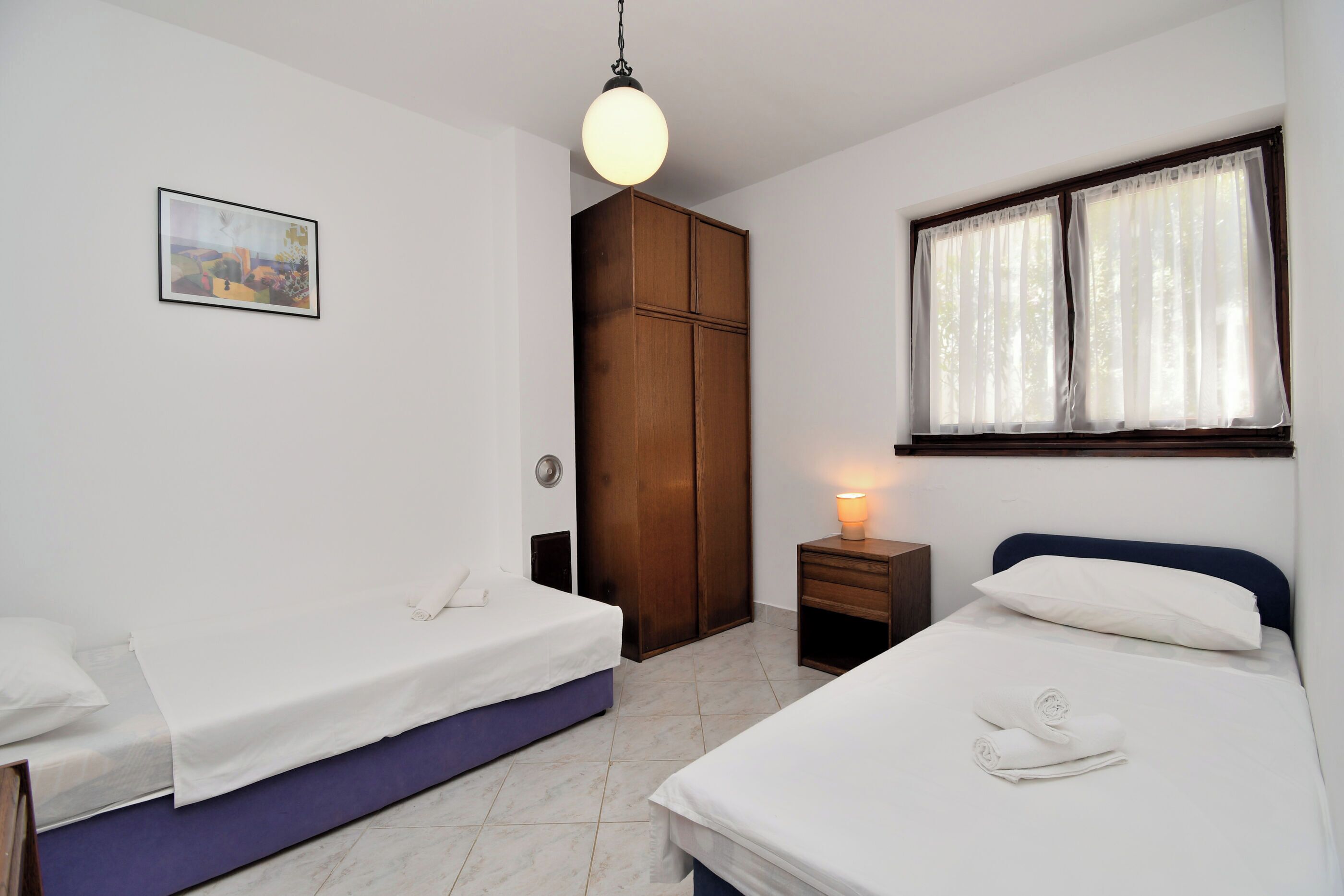 Un dormitorio con dos camas, un armario de madera, una mesita de noche con lámpara y una ventana con cortinas.