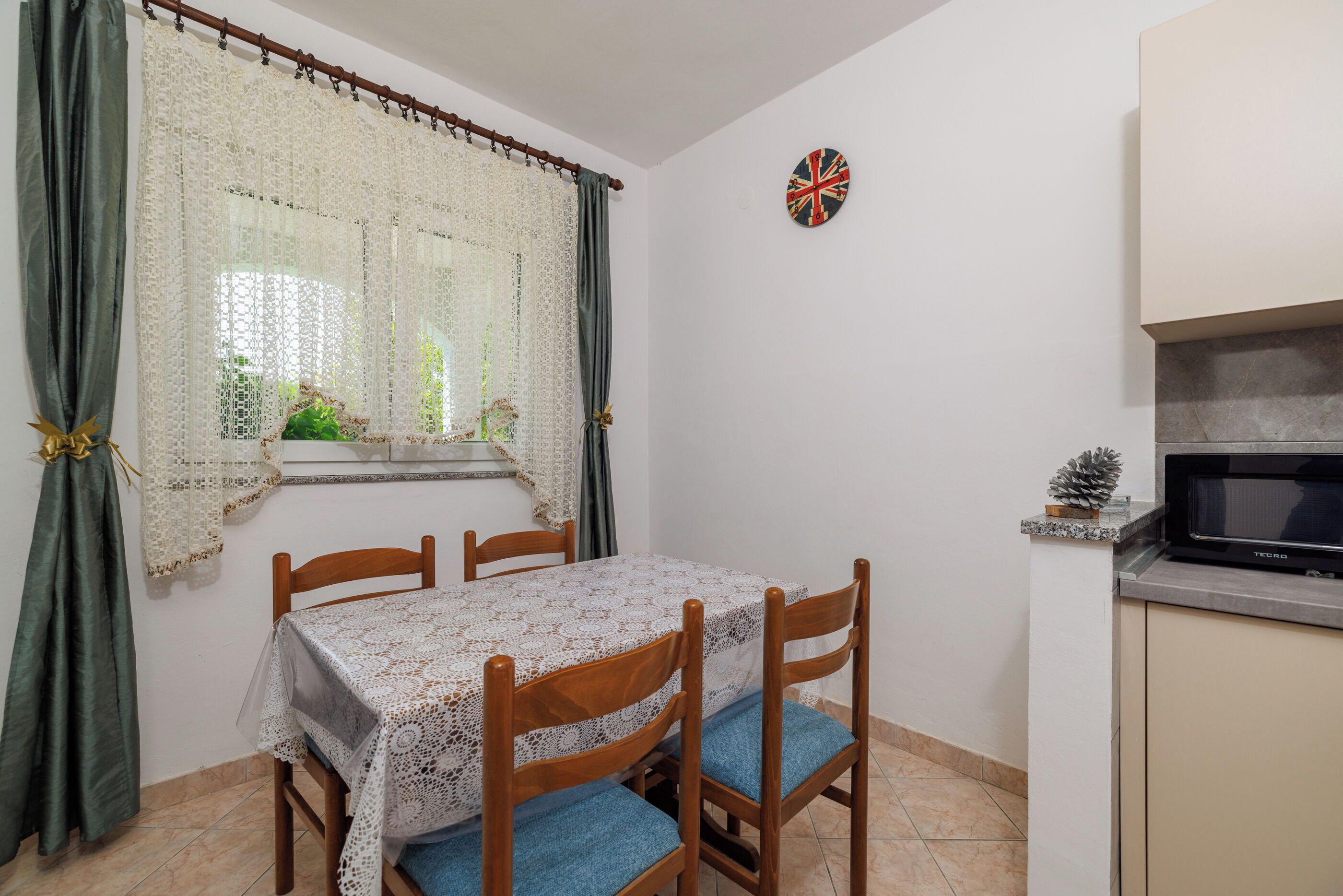 A dining area with a table covered in lace, wooden chairs with blue cushions, a window with lace curtains, a dartboard, and a microwave on the counter.