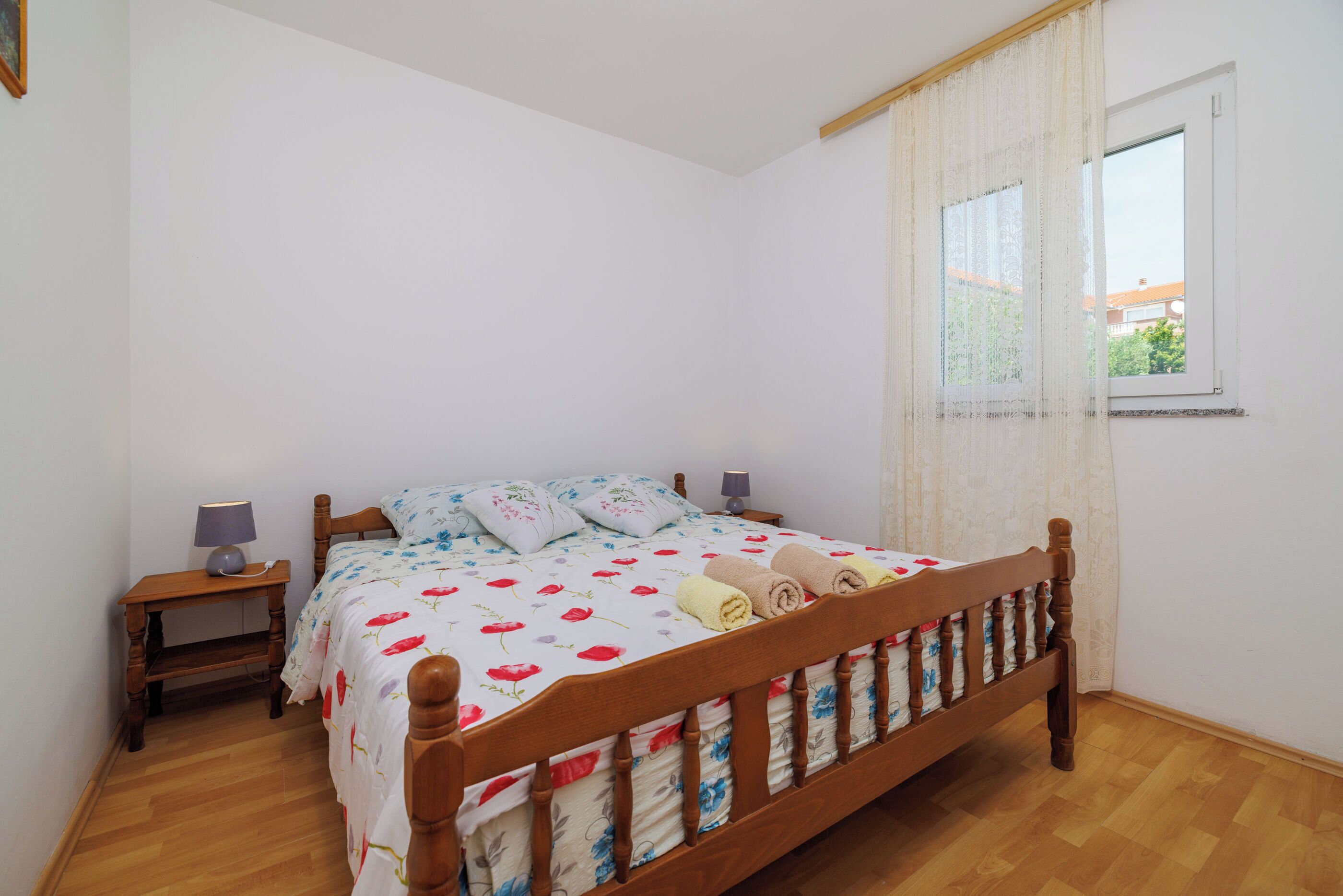 A bedroom with a wooden bed, white walls, a window with curtains, and a nightstand with a lamp.