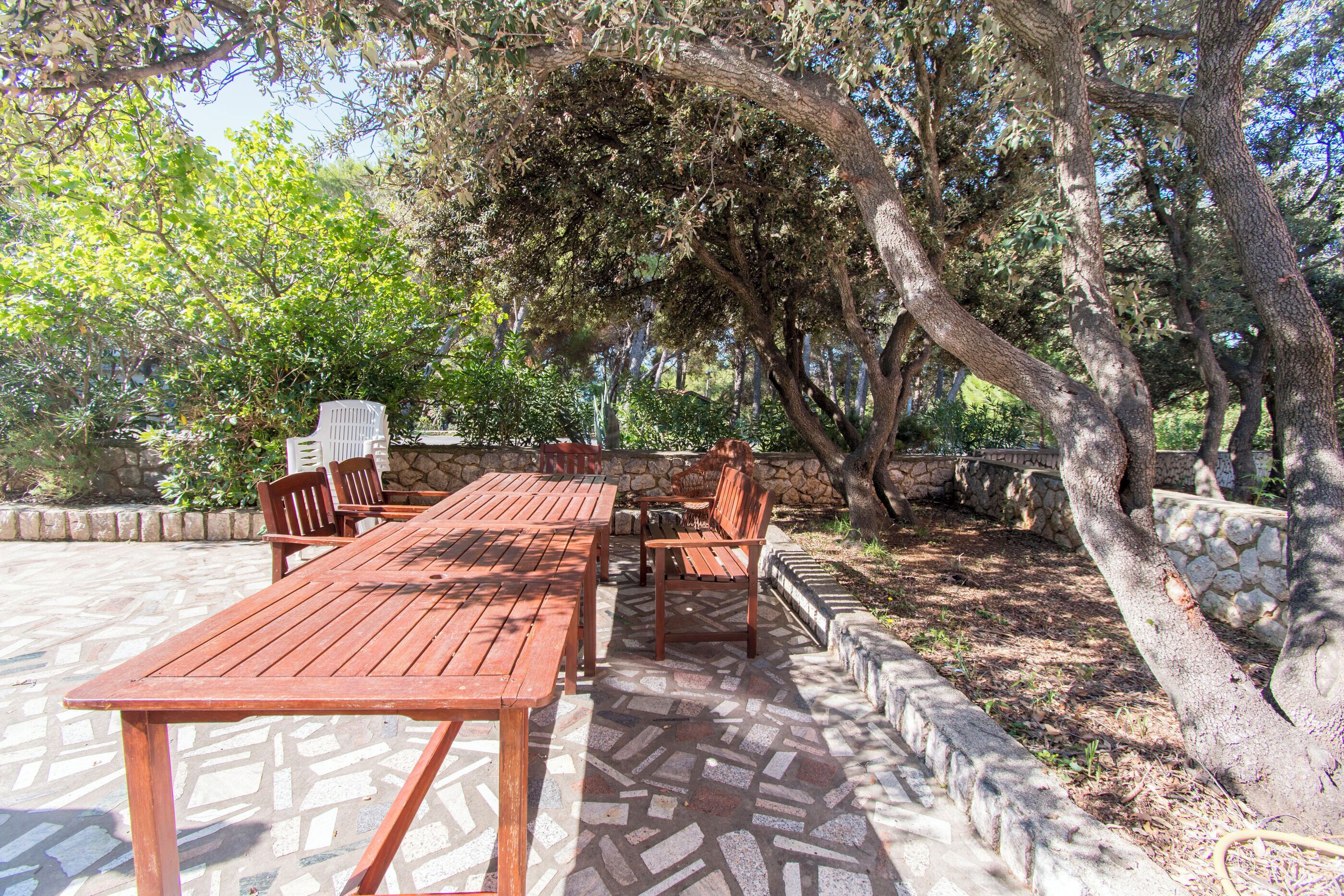 An outdoor patio with a wooden table and chairs, surrounded by stone walls and trees.