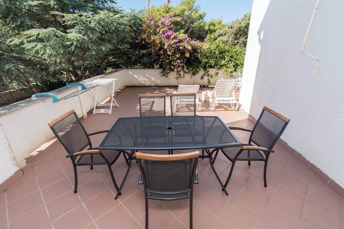 An outdoor patio with a glass-top table and four chairs, surrounded by a white wall and a garden with flowering plants.