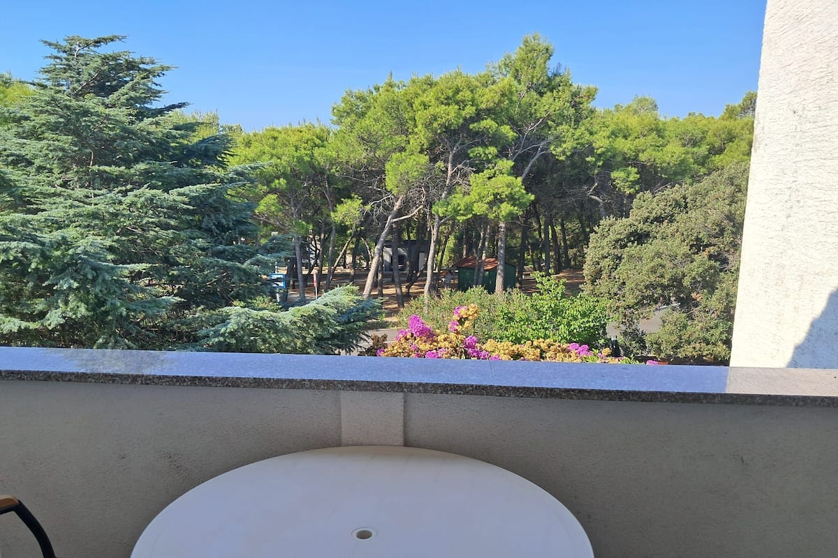 A balcony with a round table and chairs, overlooking a garden with various trees and flowering plants.