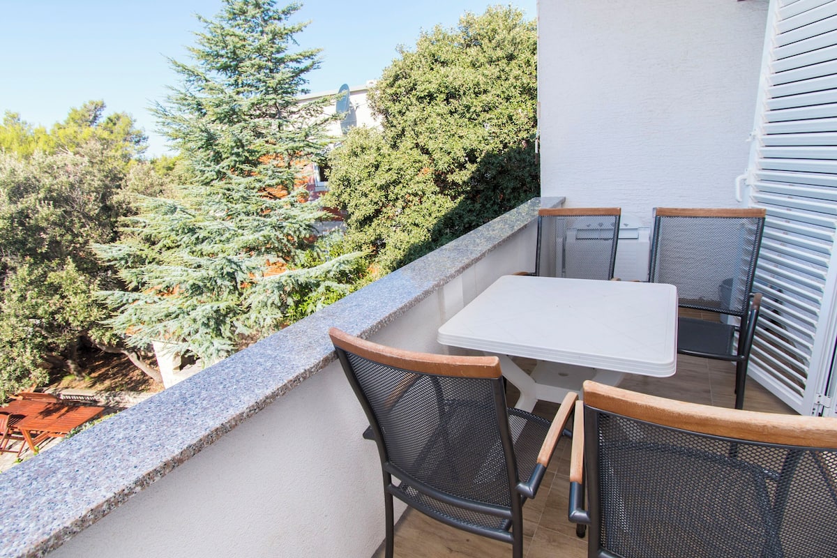 A balcony with a white table and two chairs, overlooking a garden with trees and a building in the background.