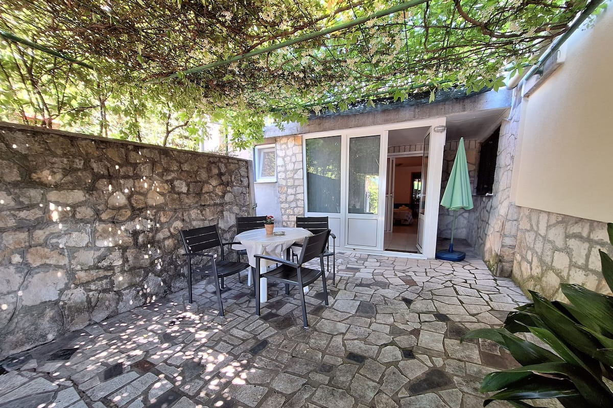 Une terrasse en pierre avec une table et des chaises, un mur en pierre et un espace couvert agrémenté d’un parasol vert.