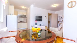 A modern living room with a glass-top dining table, white sofas, a flat-screen TV, and a kitchen in the background. - Apartment Tino (Split)