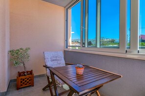 Un balcón con una mesa de madera, una silla, una planta en maceta y vistas a edificios y montañas a través de amplios ventanales.