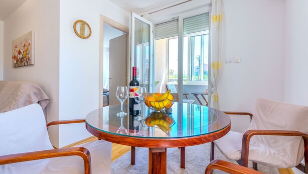 A round glass table with a wooden base, surrounded by two white cushioned chairs. A bowl of fruit and a bottle of wine are on the table. - Apartment Tino (Split)