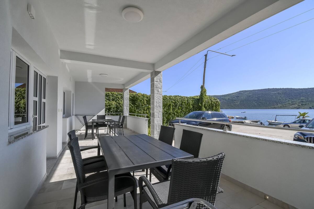 A covered outdoor patio with a dining table and chairs, overlooking a parking area and a body of water.