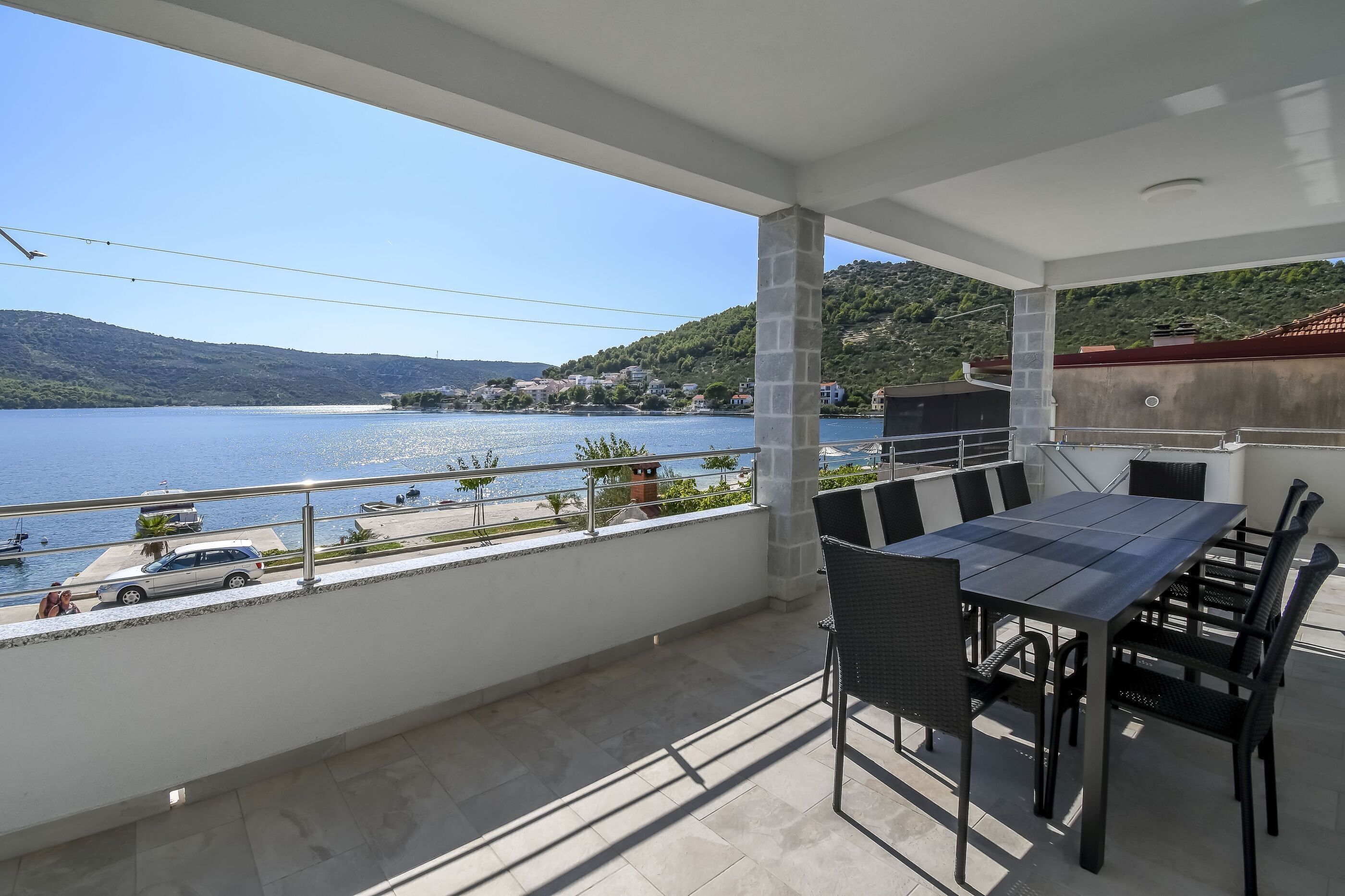 Un balcon avec une table et des chaises, donnant sur un lac où l’on aperçoit des bateaux et une voiture sur la rive.