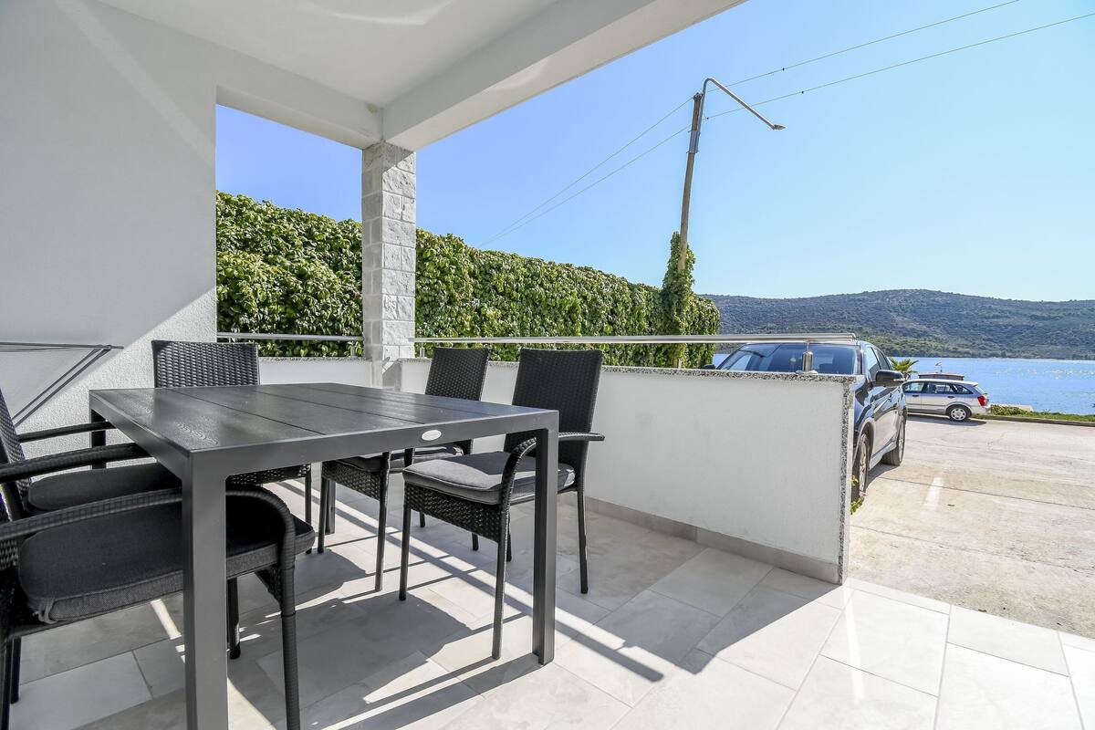 A balcony with a table and chairs, a car parked outside, and a view of the sea and hills.