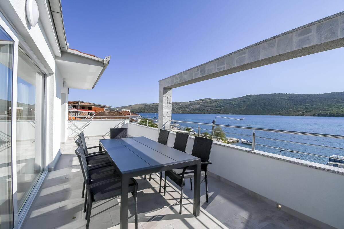 A balcony with a table and chairs, overlooking a body of water and hills.