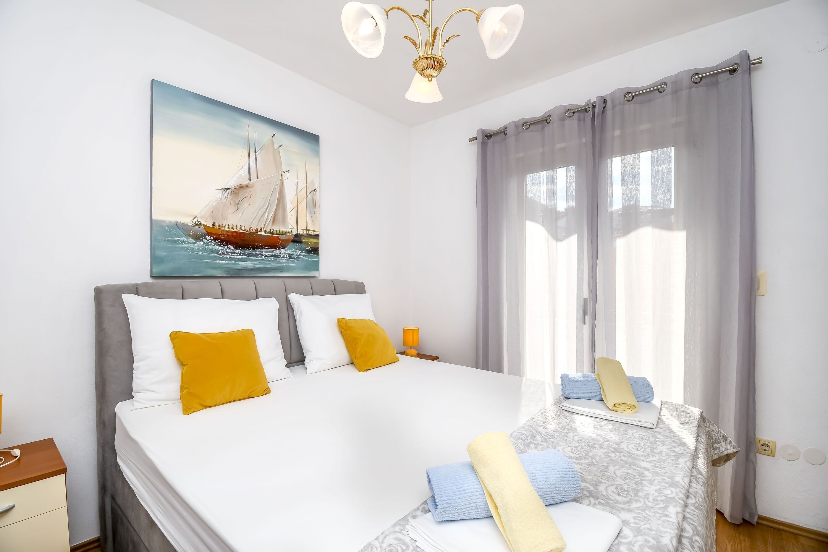 A neatly made bed with pillows, a bedside table with a lamp, and a painting of a sailboat on the wall.