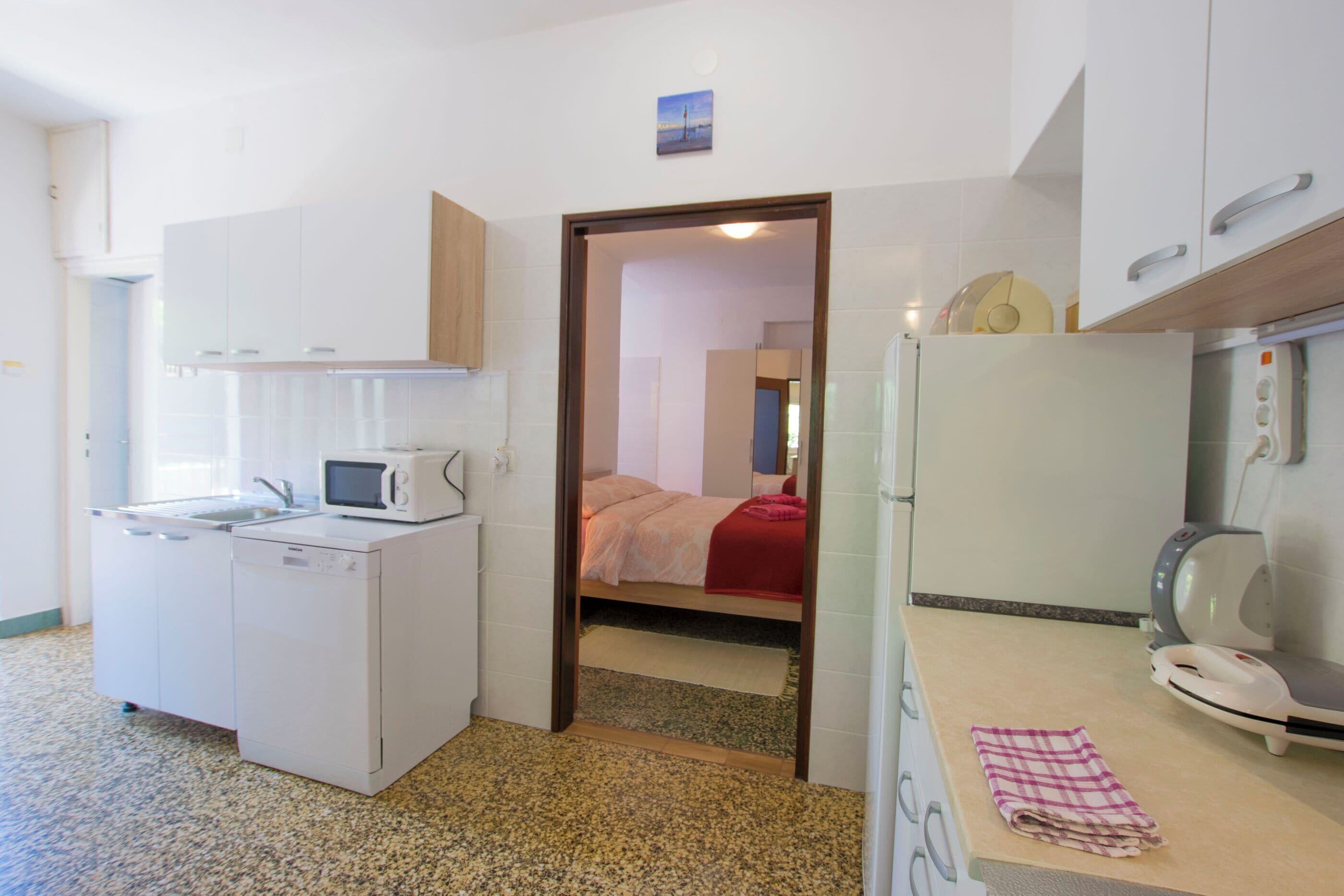Cocina compacta con microondas, lavavajillas y tabla de planchar. Se ve una cama a través de una puerta entreabierta al fondo.