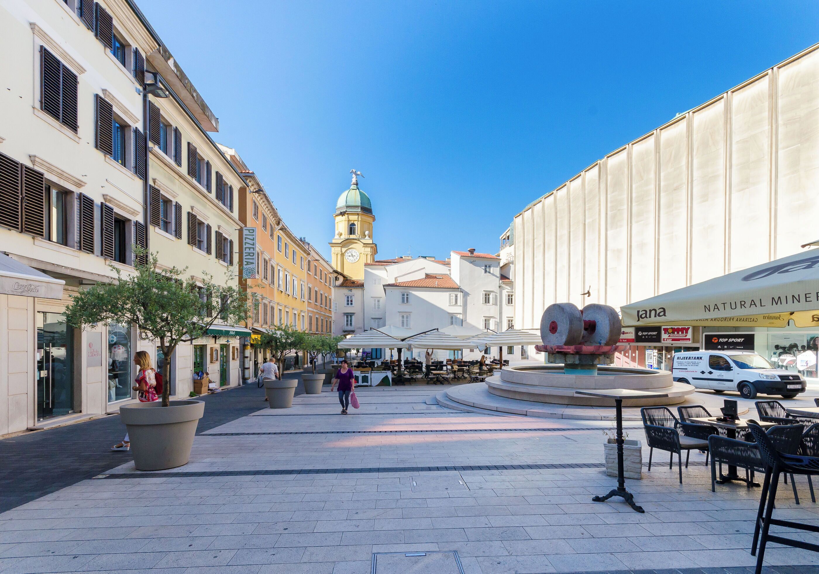 A public square with a fountain, outdoor seating, and a clock tower in the background.