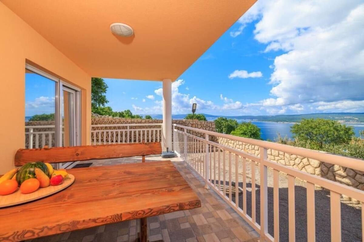 Ein Balkon mit Holztisch und Bank, eine Steinmauer und Blick auf einen See und Berge.