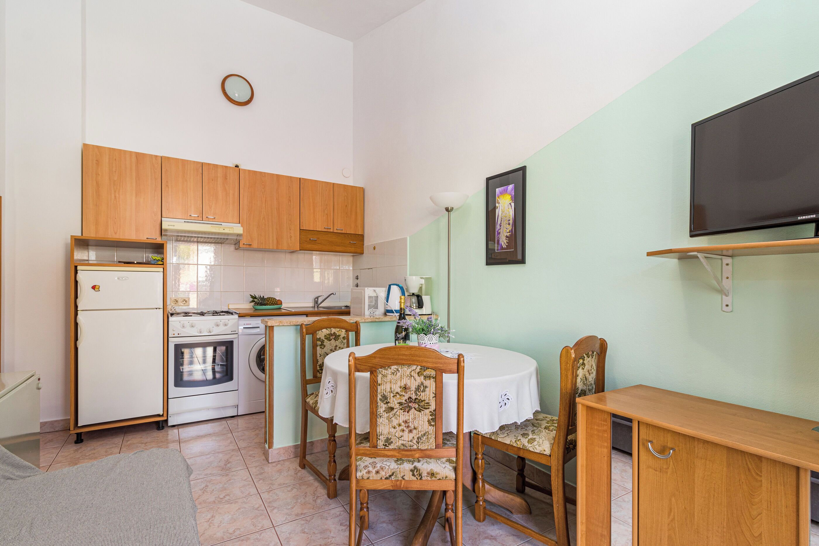 Une cuisine compacte avec des meubles en bois, un réfrigérateur, un four et un lave-linge. On y trouve une table à manger avec des chaises et un téléviseur à écran plat fixé au mur.