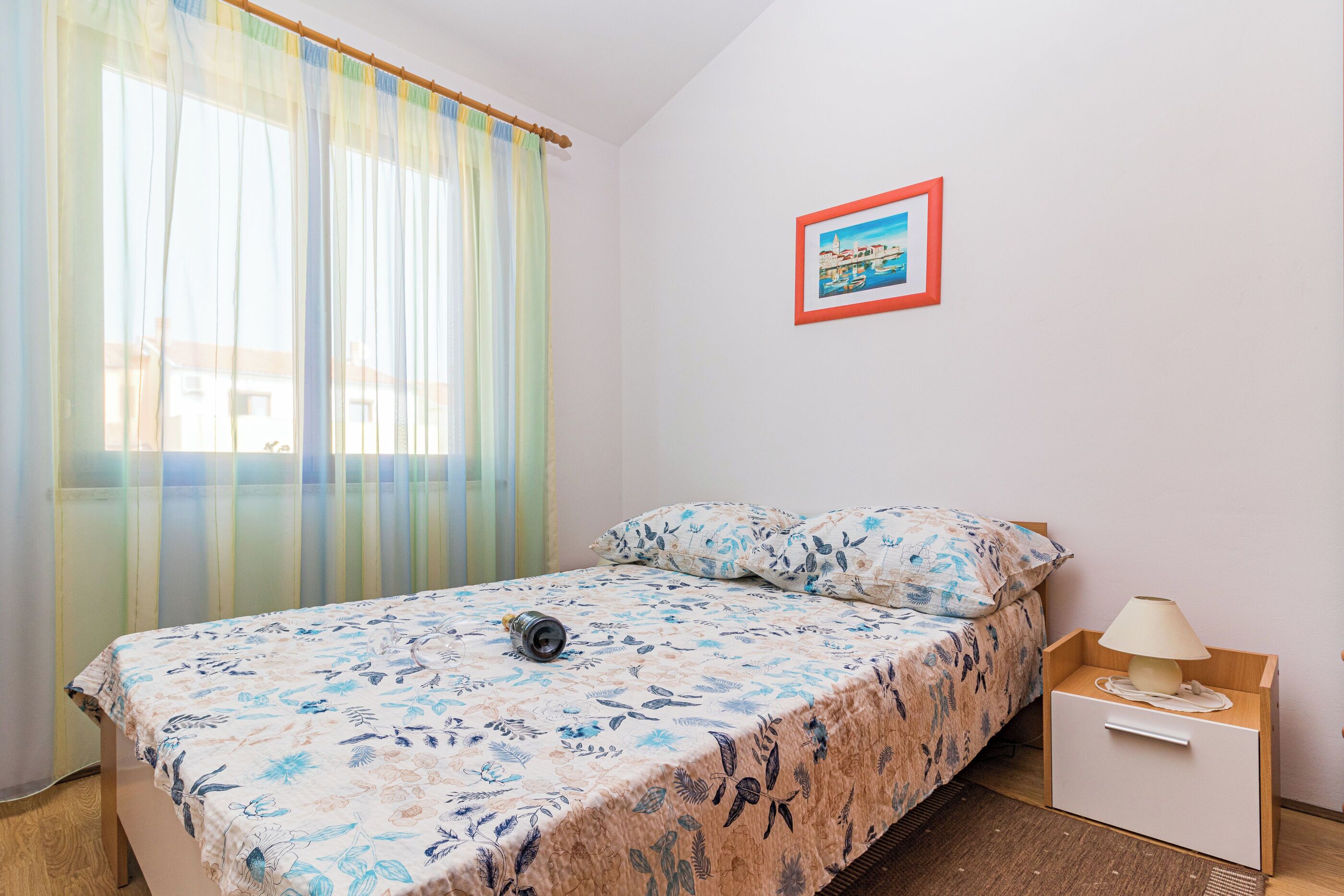 Una camera da letto con un letto, un comodino, una lampada, una finestra con tende leggere e un quadro appeso al muro.