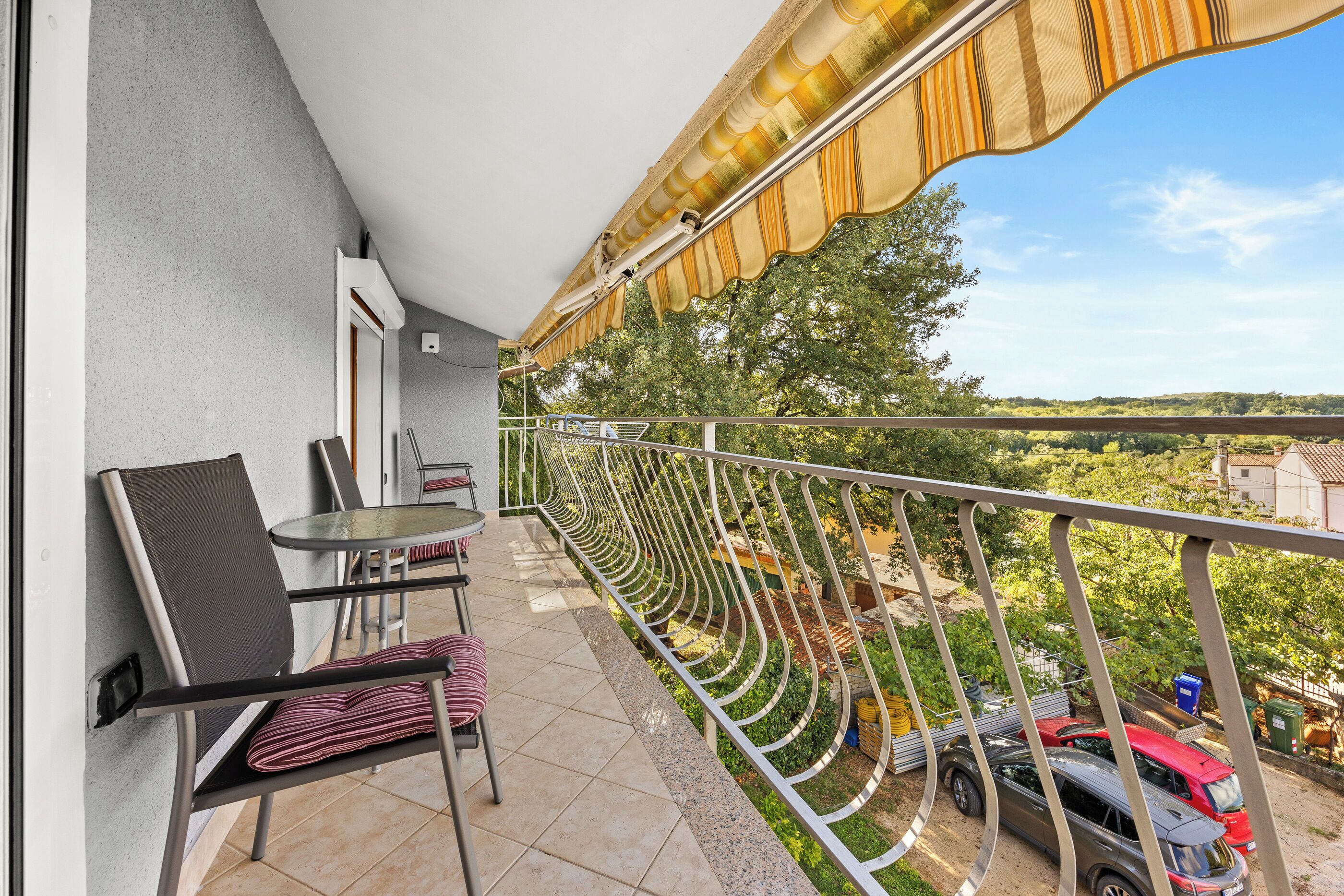 Een balkon met een tafel en stoelen, uitzicht op de omliggende begroeiing, en geparkeerde auto's beneden.