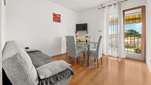 Un salon moderne avec un canapé gris, une table à manger avec des chaises, un téléviseur à écran plat fixé au mur et une grande fenêtre avec des rideaux.