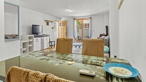 A modern living room with a glass-top dining table, two chairs, a television, and a kitchen area with cabinets and a sink. - Apartment Tin (Medulin)