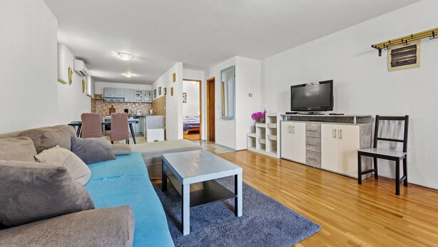 A modern living room with a sofa, a coffee table, and a TV. There is a kitchen area in the background with cabinets and a dining table. - Apartment Tin (Medulin)