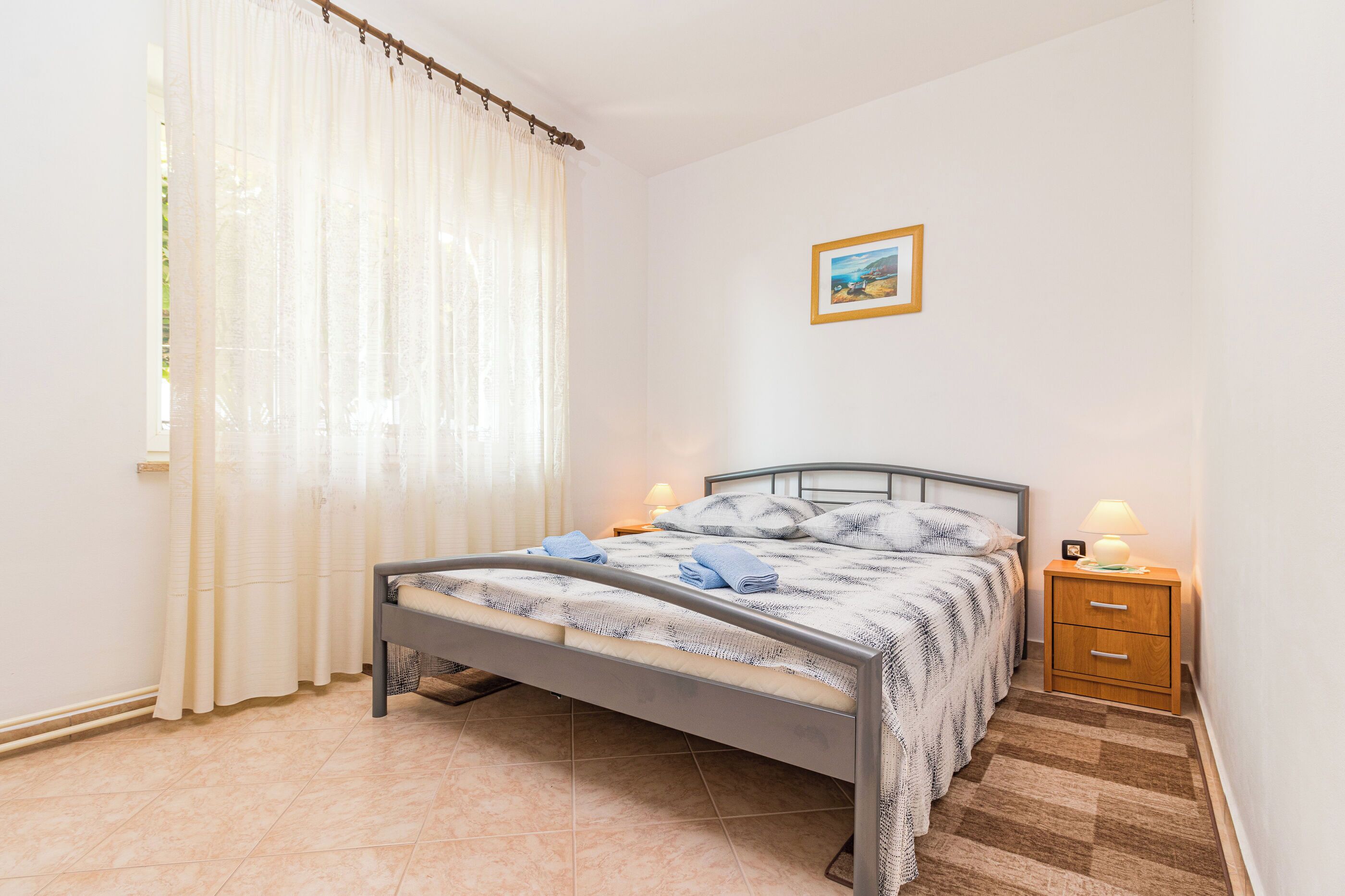 A bedroom with a bed, a nightstand, a lamp, a picture on the wall, and a window with curtains.