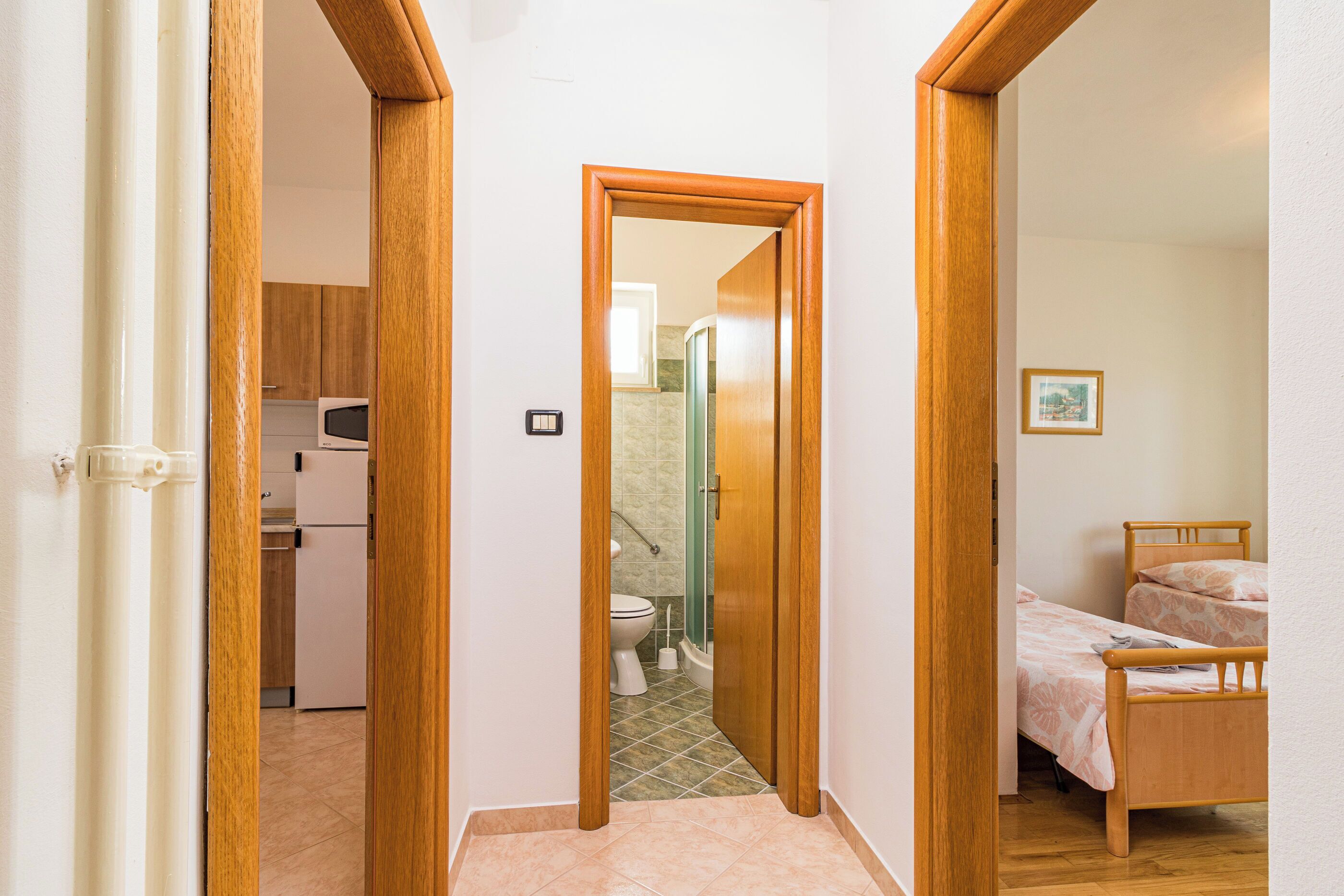 Ein schmaler Flur führt zu einem Badezimmer mit Toilette und Dusche, einem Bett mit Holzrahmen sowie einer Küchenzeile mit Kühlschrank und Mikrowelle.