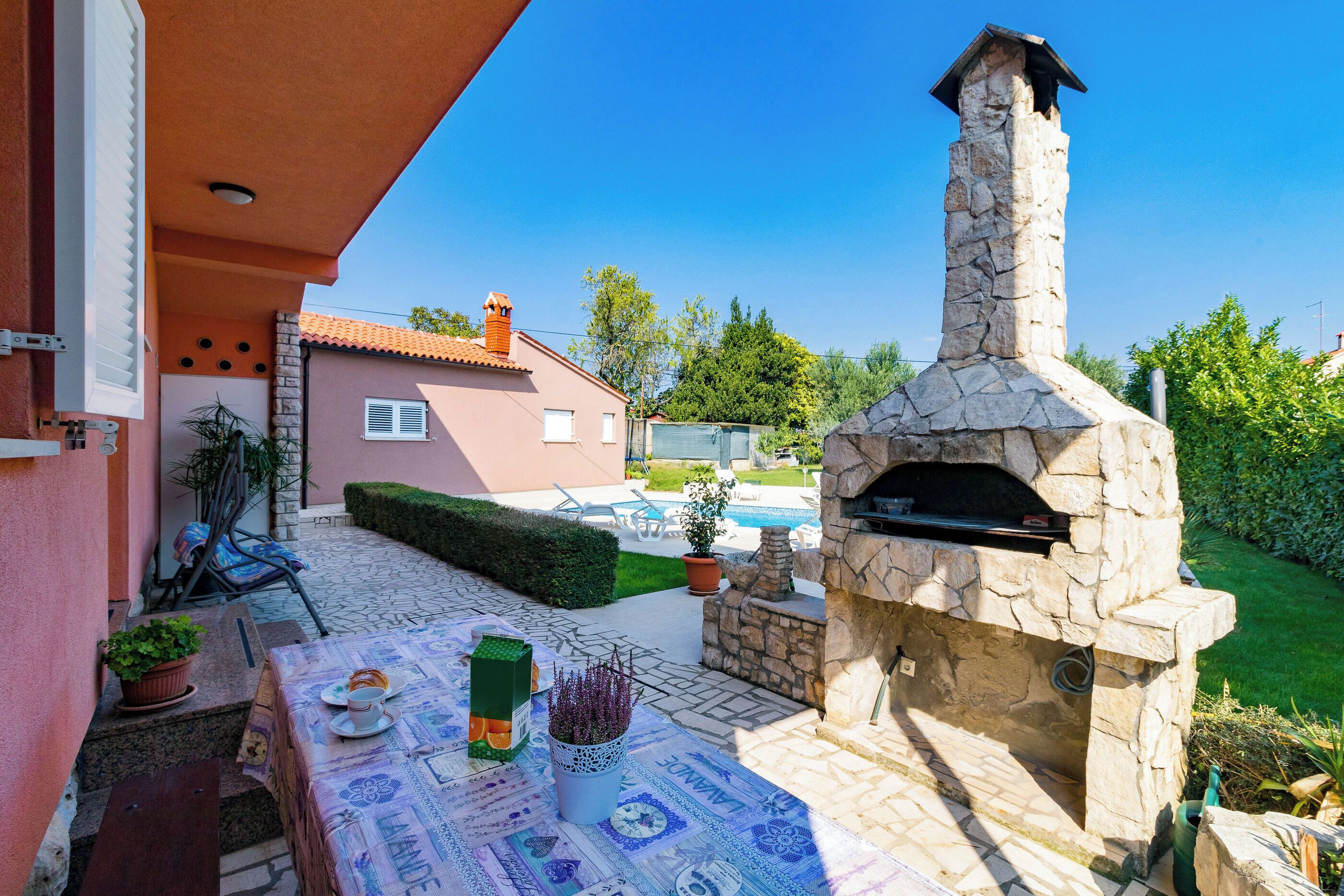Une terrasse extérieure avec un four à pizza en pierre, une table recouverte d’une nappe et une piscine en arrière-plan.