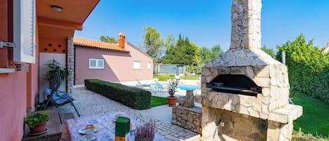 Une terrasse extérieure avec un four à pizza en pierre, une table recouverte d’une nappe et une piscine en arrière-plan.