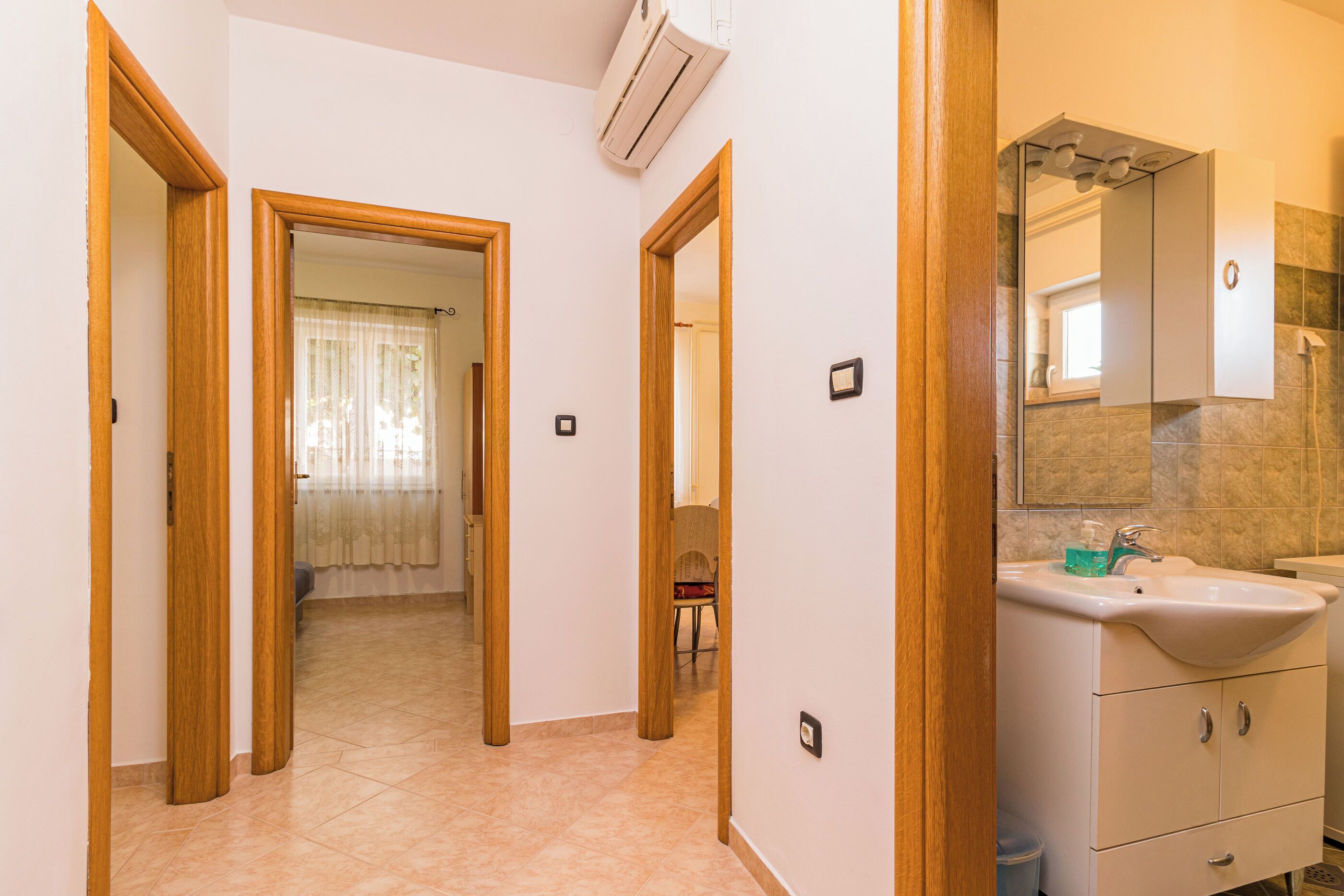 Un pasillo con puertas de madera que dan a una habitación con cama y ventana con cortinas. Hay un baño con lavamanos y espejo.