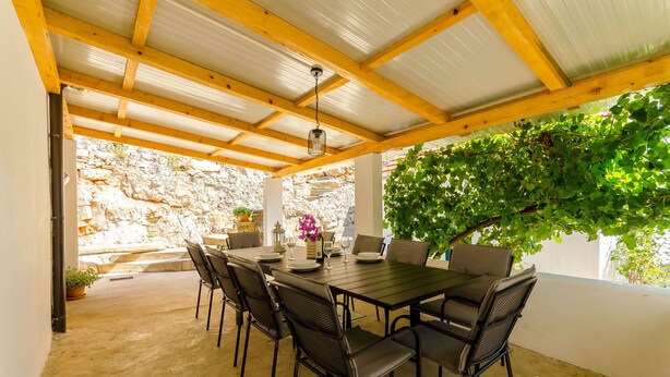 An outdoor covered patio with a table set for dining, surrounded by chairs, and a stone wall in the background. - House Mateljan (Jelsa)