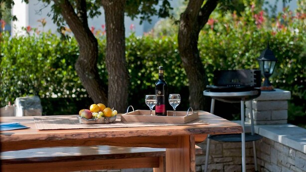 Uma área de refeições ao ar livre com mesa de madeira, uma garrafa de vinho e um tabuleiro com frutas.