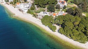 An aerial view of a coastal area with a beach, houses, and dense greenery. - Apartments Kovami (Podgora)