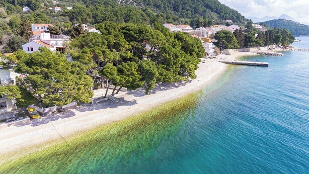 A coastal area with a beach, clear water, and residential buildings. - Apartments Kovami (Podgora)