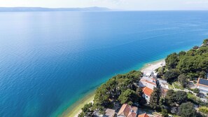 An aerial view of a coastal area with clear blue waters, a sandy beach, and a cluster of buildings with red-tiled roofs. - Apartments Kovami (Podgora)