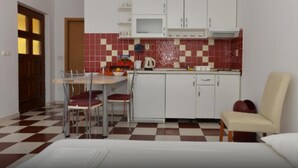A compact kitchen with white cabinets, a red and white checkered floor, a dining table with chairs, and a small sofa. - Apartments Kovami (Podgora)