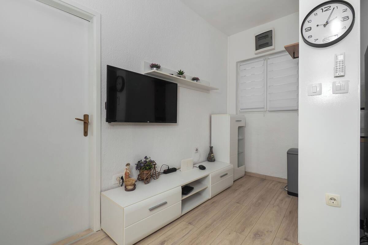 A room with a TV mounted on the wall, a white cabinet, a small shelf with plants, a clock, and a door.