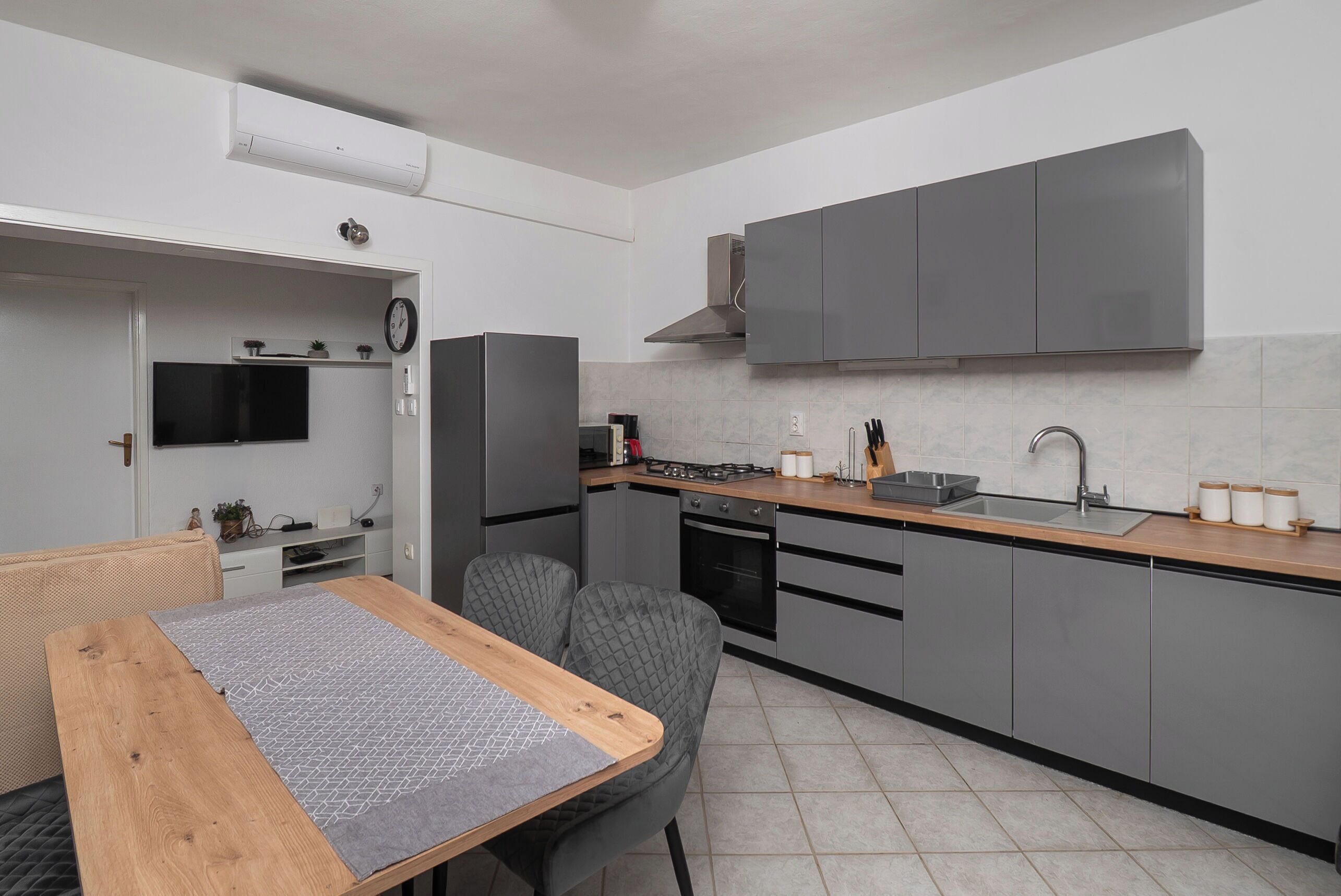 A compact kitchen with grey cabinets, a wooden countertop, a built-in stove, and a wall-mounted TV.
