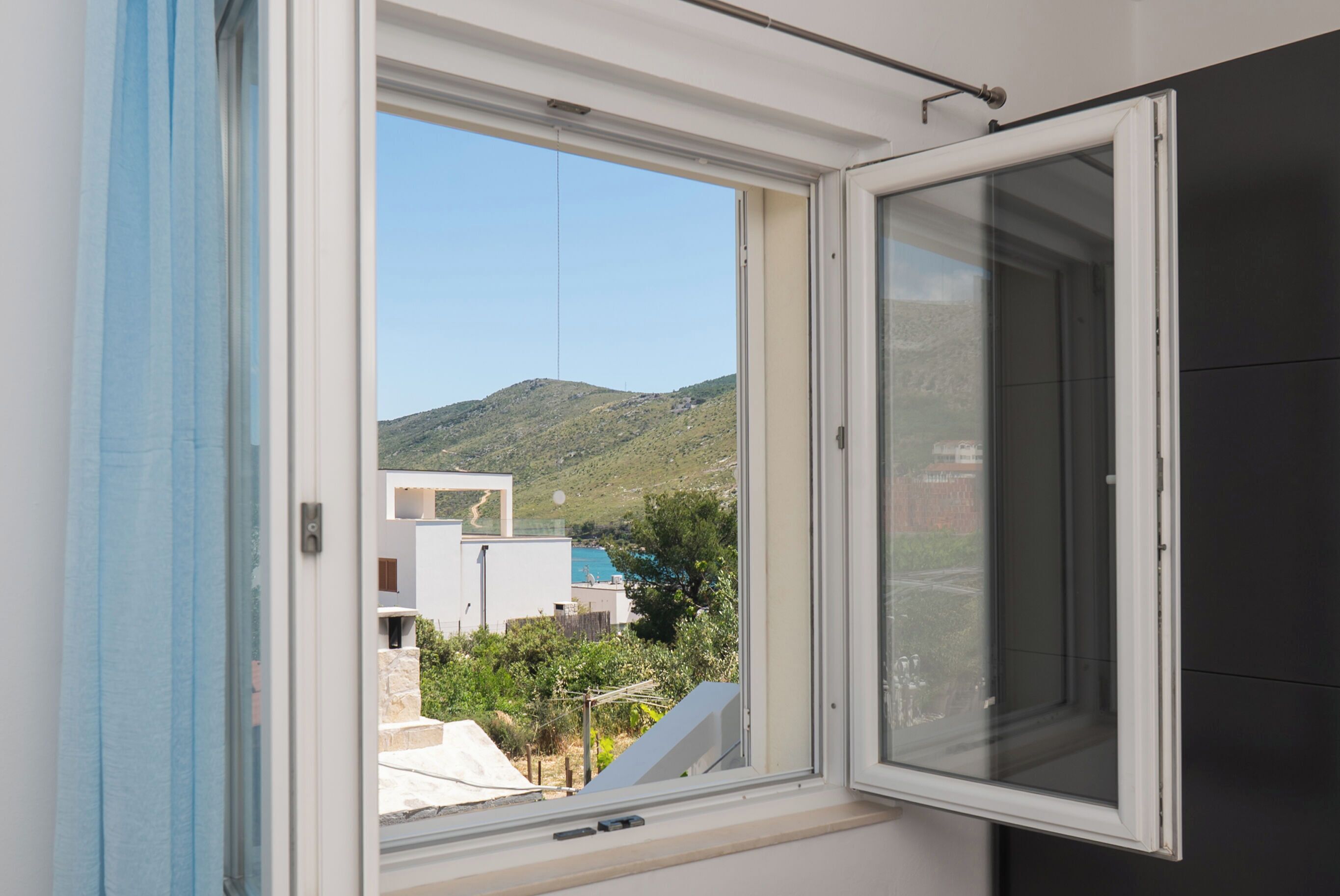 A room with a large window, a view of a hillside, and a glimpse of buildings outside.