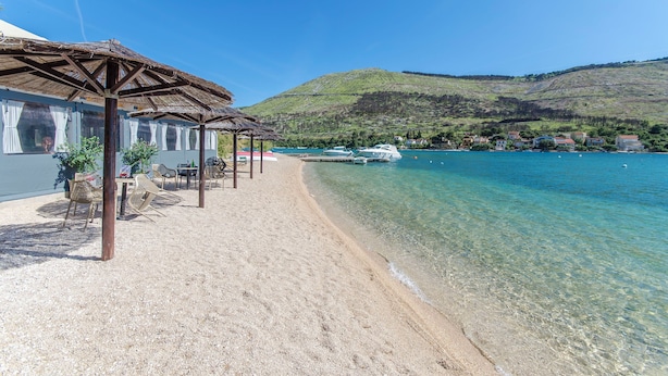 A beach with clear water, a wooden structure with chairs, and boats in the background. - Apartments Jadranka (Sibenik)
