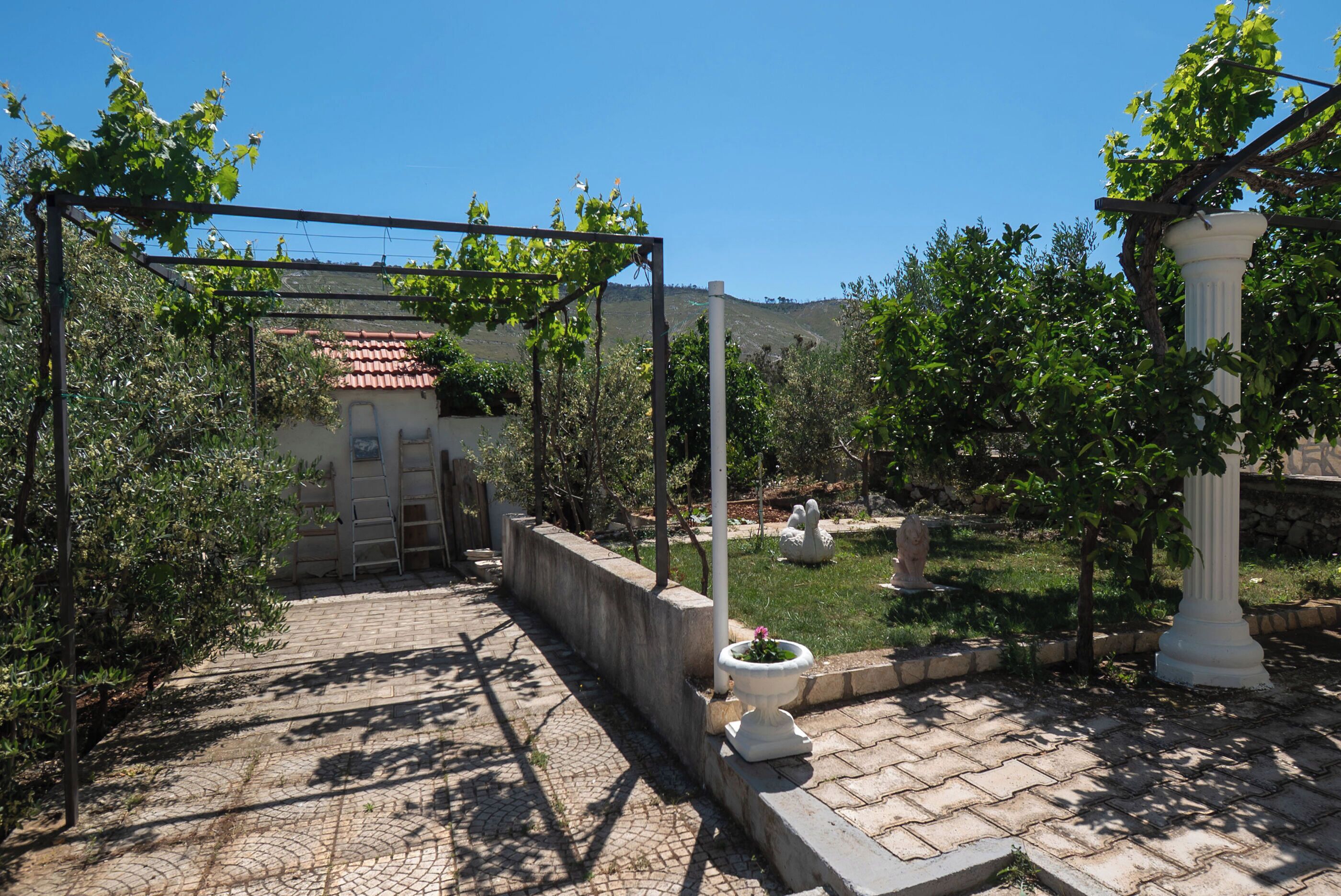 Un chemin pavé mène à un bâtiment dont le toit est recouvert de tuiles rouges, et qui est entouré de verdure et d’une structure en forme de pergola.