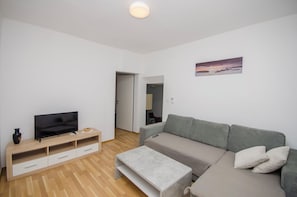 A modern living room with a flat-screen TV on a wooden TV stand, a grey sofa, and a small coffee table. - Apartments Mirjana (Nin)