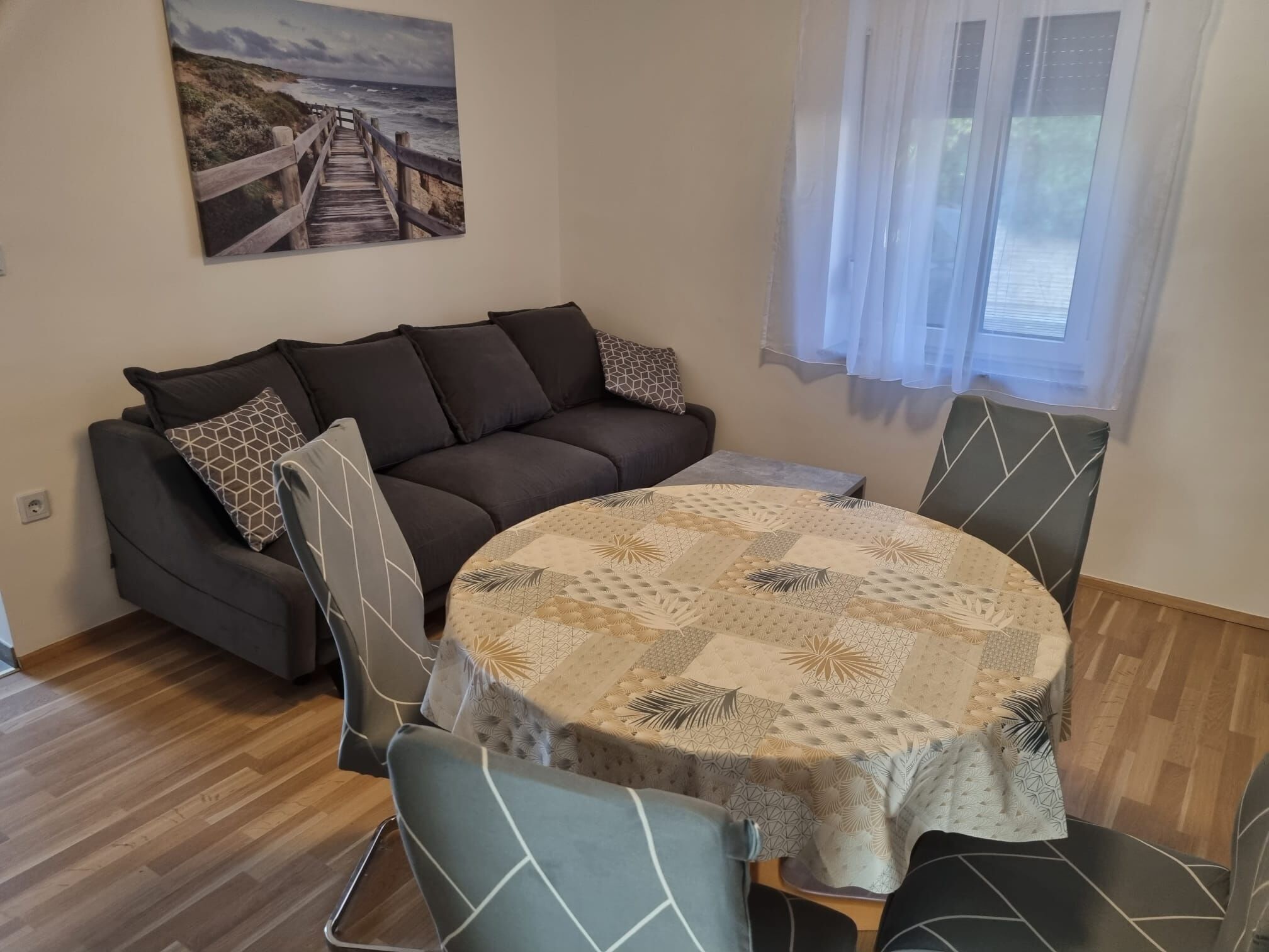 Ein gemütlicher Wohnbereich mit einem dunkelbraunen Sofa, gemusterten Kissen und einem runden Tisch mit einer gesteppten Tischdecke. An der Wand hängt ein Bild von einem Steg und dem Meer.