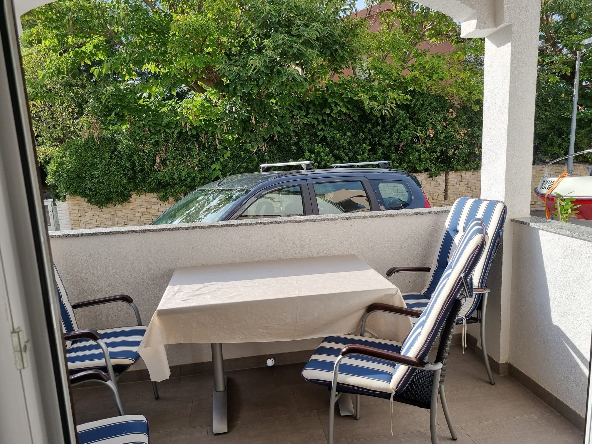 A balcony with a table and striped chairs, a parked car, and lush greenery in the background.