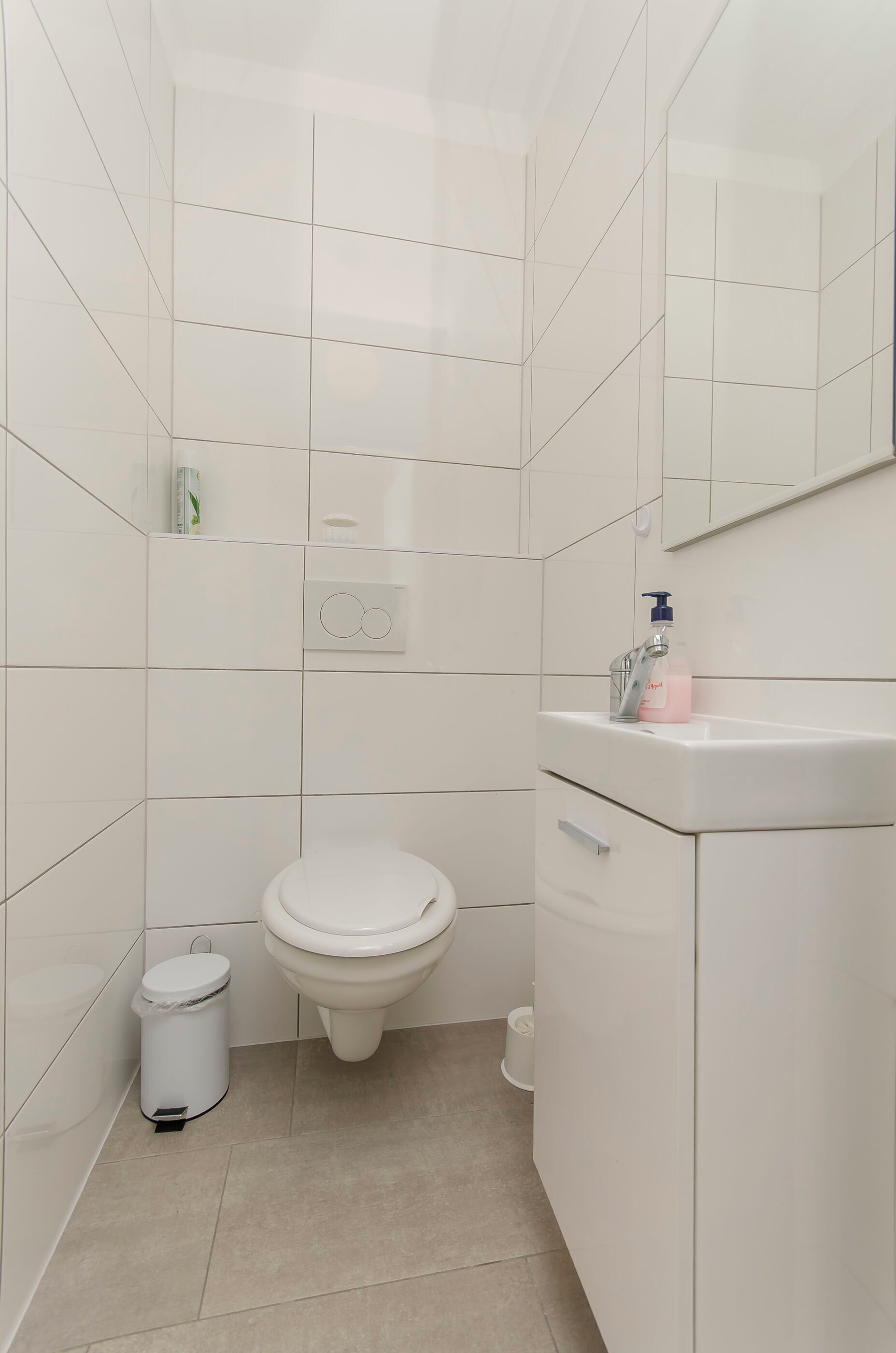 A small, modern bathroom with white tiles, a toilet, a sink with a mirror, and a trash bin.